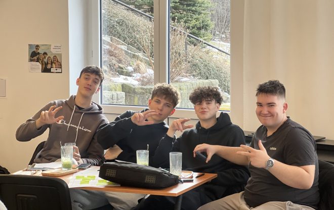 Skupina čtyř studentů u stolu během podnikatelského workshopu; sklenice s nápoji, poznámkové lístky a okno s venkovní zelení v pozadí.
